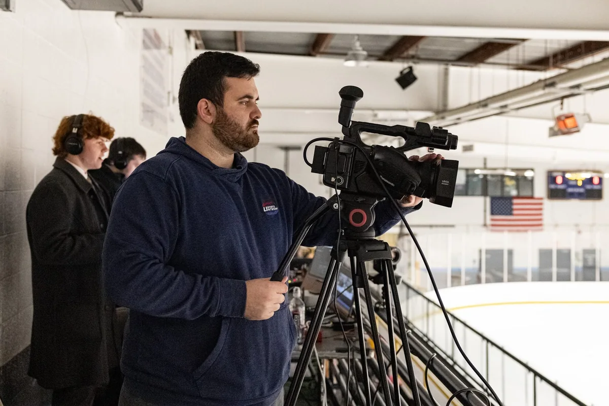An LI Sports Network camera operator running a broadcast from the rink, with the announcer calling the game behind him